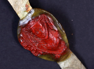 A red wax seal in the shape of a veiled woman's face on a strip of leathery material.