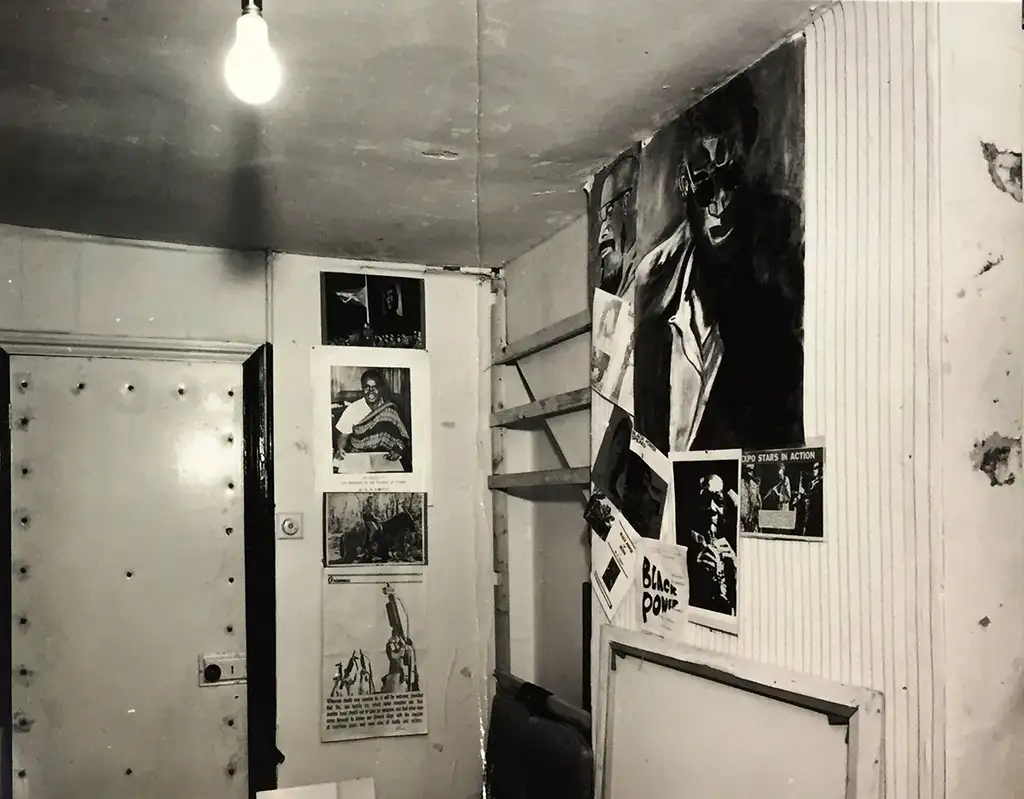 A small, cramped room with no windows. On the walls are posters and drawings.