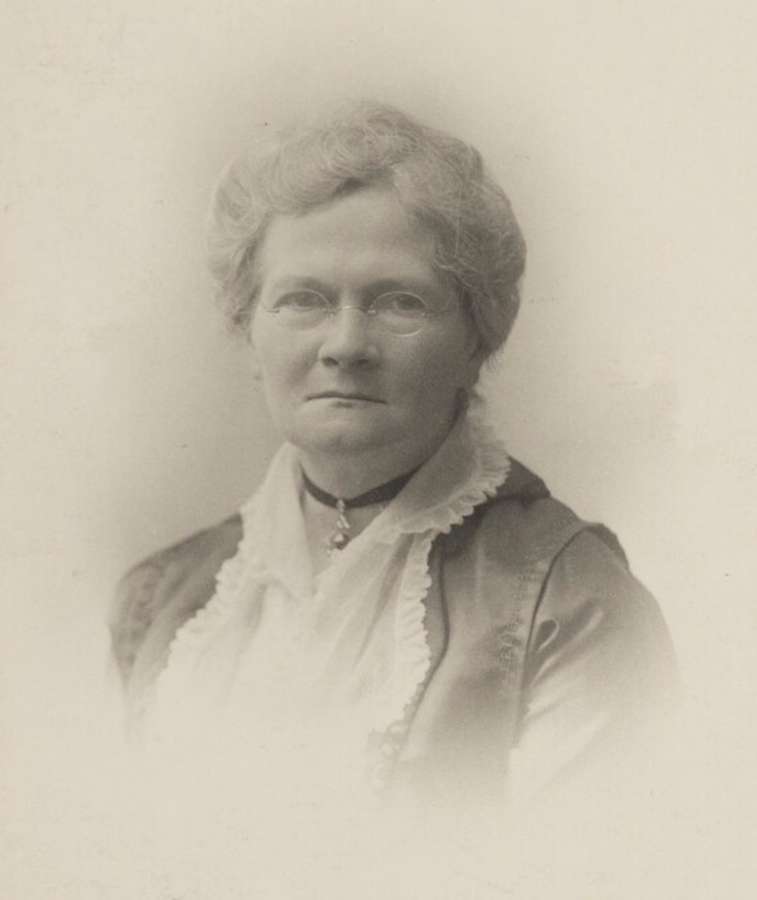 A middle aged woman in smart clothes wearing small round glasses. Her expression is thoughtful.