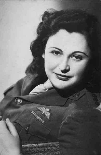 A woman in military uniform with shoulder length dark hair looks over her shoulder at the camera