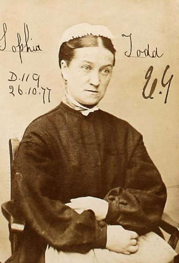 A woman in a black dress with white cap sits with her arms folded in her lap