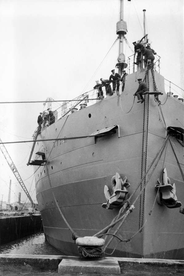 Black and white photograph of the front of a docked ship, with around ten crew working on it.