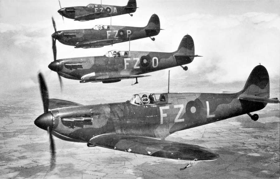 Black and white photograph of four Spitfire aircraft flying in a horizontal line over fields.