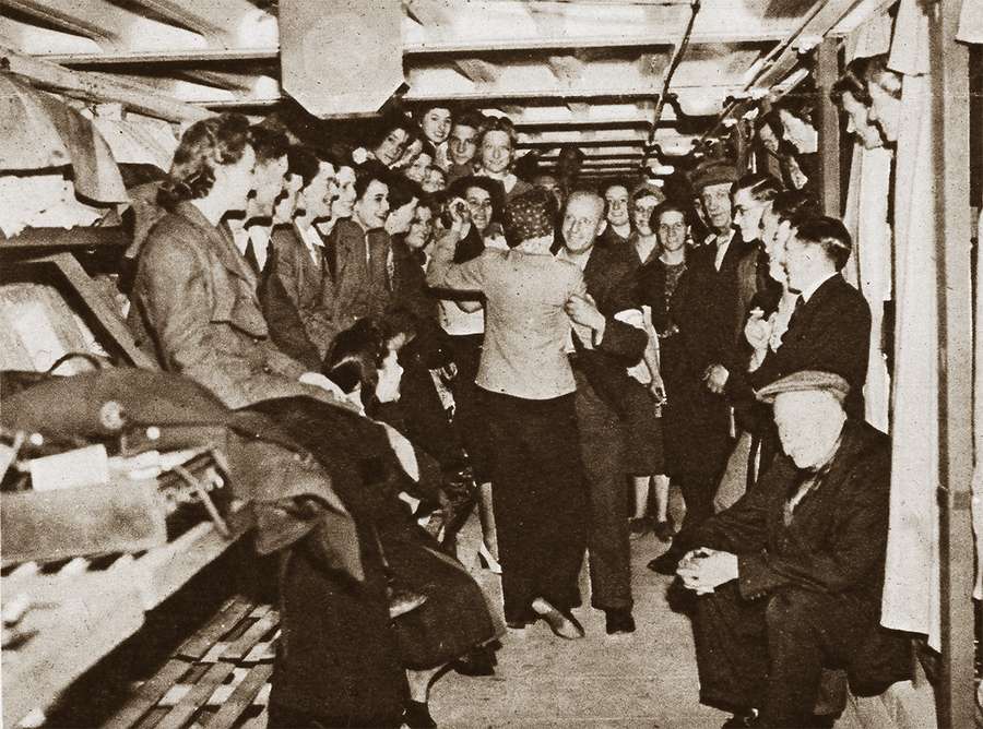 Dozens of people, all smiling, squeezed into a corridor-like shelter as two do ballroom dancing.