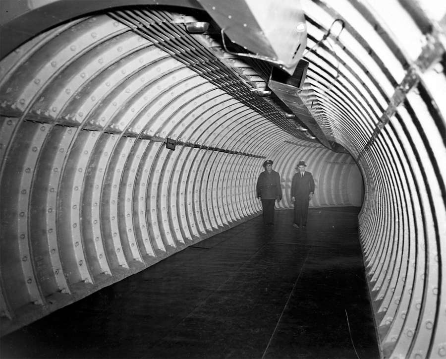 A man in military uniform walks beside a man in a suit down a curved, wide tunnel with metal ribs.