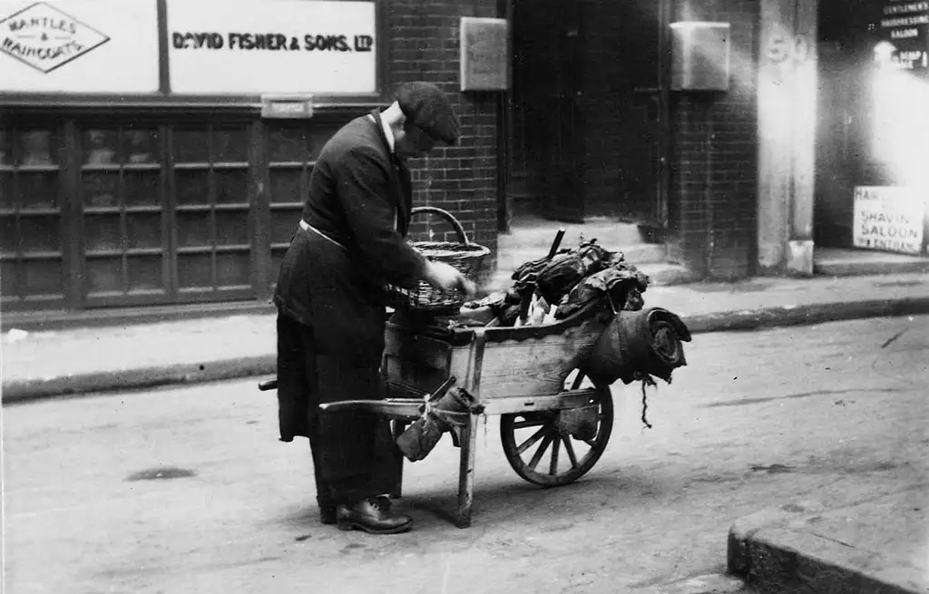 A man wearing a flat cap and holding a basket reaches into the wheelbarrow in front of him.