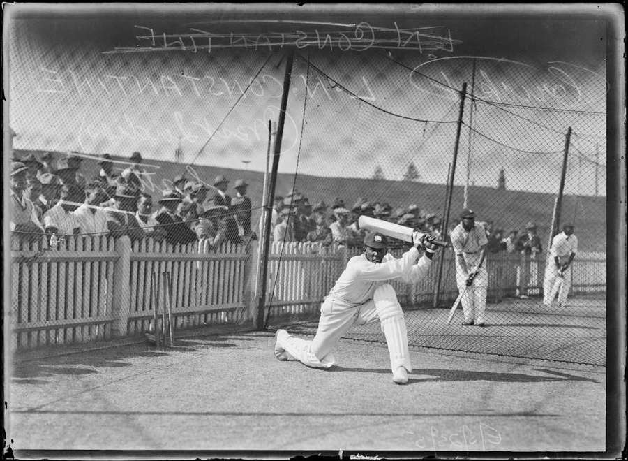 A young black man in cricketing whites photographed in the middle of hitting the ball. A crowd watch