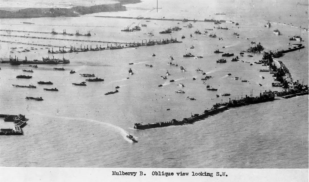 Aerial photograph showing dozens of ships, large and small, between a Mulberry harbour and cliffs.