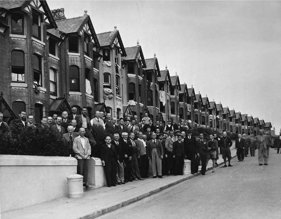 Black and white photograph showing dozens of men gathered outside a row of terraced houses.