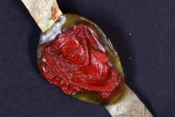 A red wax seal in the shape of a veiled woman's face on a strip of leathery material.