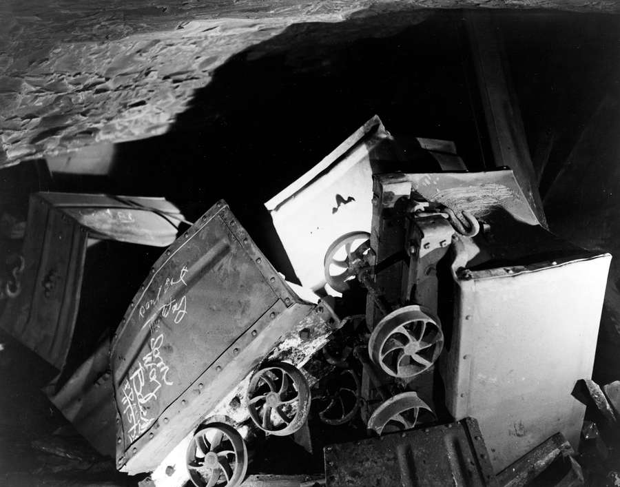 A photograph of inside a mine. There are coal trucks and rails upended. A torch light shines on them