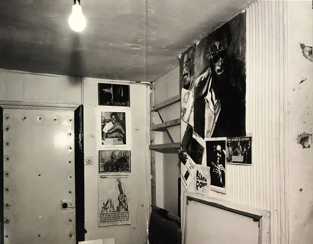 A small, cramped room with no windows. On the walls are posters and drawings.