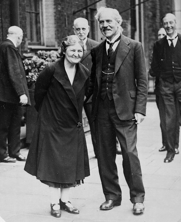 A woman in a black coat smiles at someone off camera. Next to her stands a man in a black suit