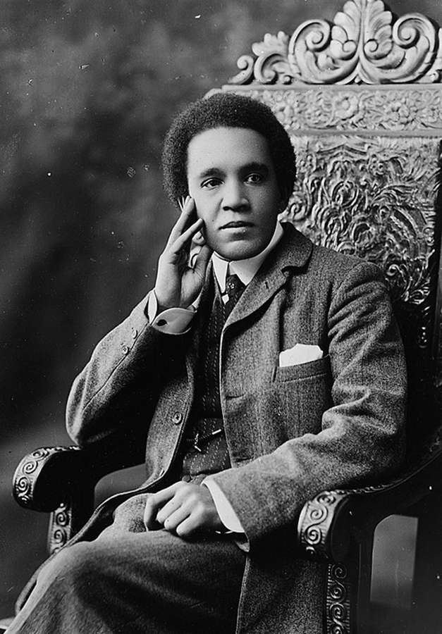 A young mixed-heritage man in a smart suit sits on an elaborately carved chair in a thoughtful pose.