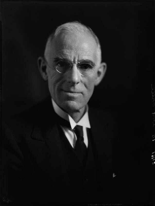 Black and white portrait photo of a smartly dressed man with short hair and glasses, smiling.