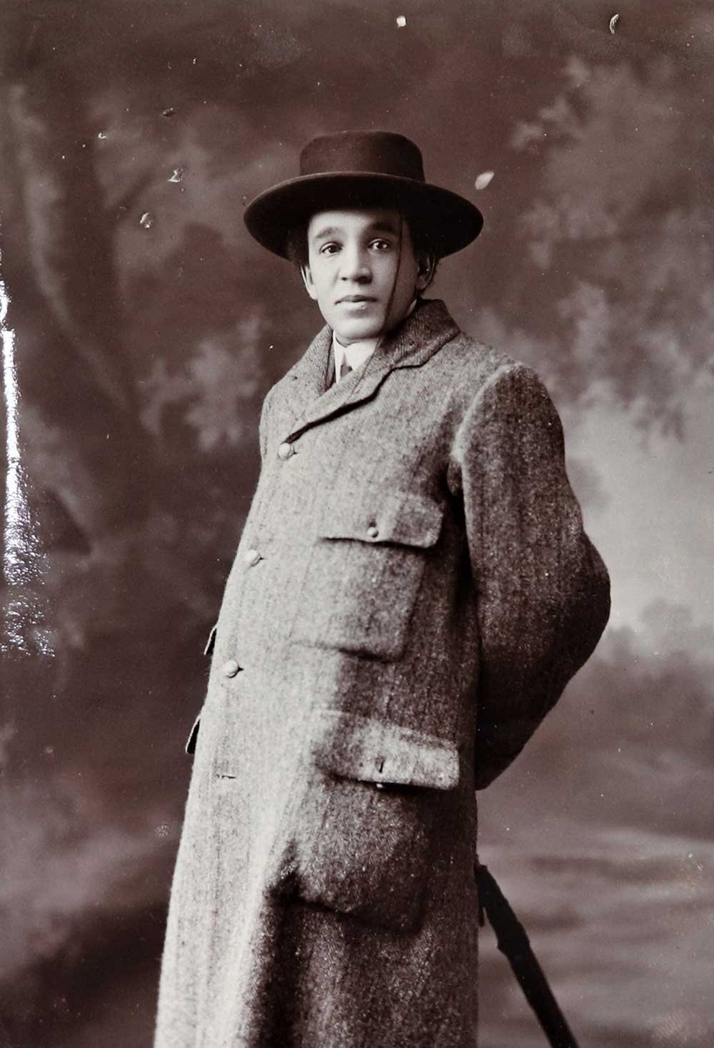 A full length shot of Coleridge-Taylor wearing an overcoat and a hat and leaning on an umbrella.