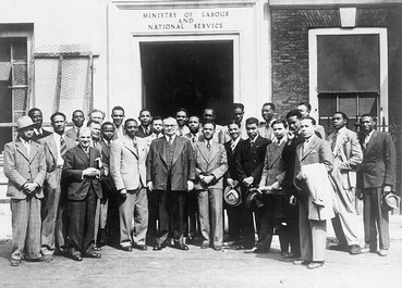 A group of 27 men in suits stand in front of a government building. Most of the men are black.