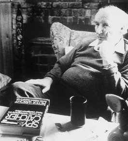 An older man sitting in an armchair with a mug and a copy of Spycatcher on the table in front.