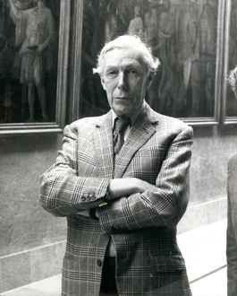 Blunt in a tweed jacket with a stern expression and his arms folded, stood next to some paintings.