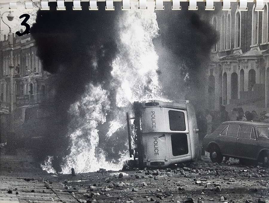 Black and white photo showing a police van on its side amid huge flames on a rubble-strewn road.