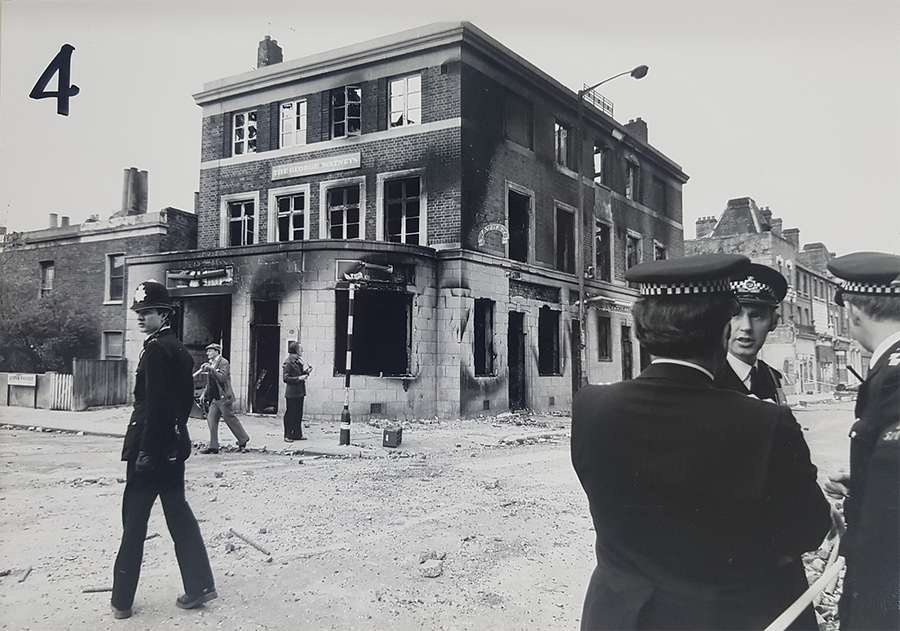 Four police officers stand outside a ruined 3-storey pub with windows smashed and many scorch marks.