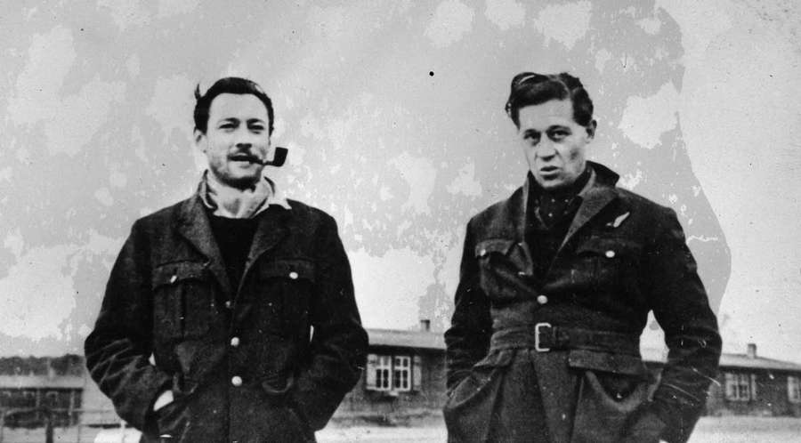 Two men standing next to each other in thick coats, one smoking a pipe, in front of some huts.