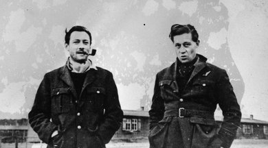 Two men standing next to each other in thick coats, one smoking a pipe, in front of some huts.