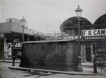 Black and white photo of a public toilet building between lampposts, with roads either side.