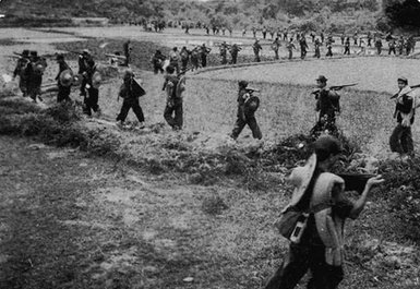A photograph of a military group marching one by one in a line through fields.