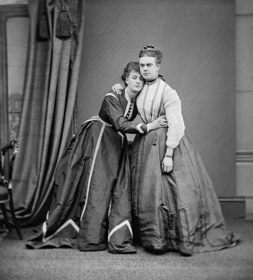 Two people in Victorian dresses and feminine hairstyles. They affectionately lean on each other.