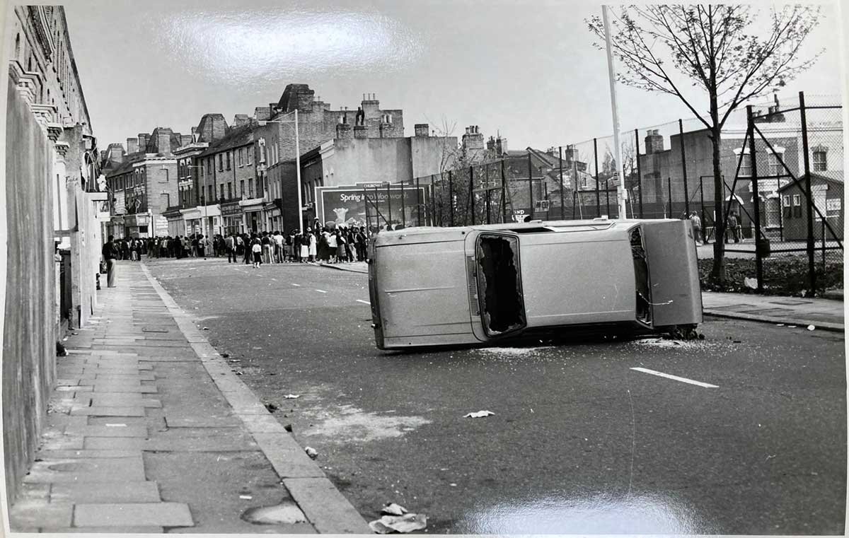 Black and white photo of a car with smashed windows lying on its side in the street.