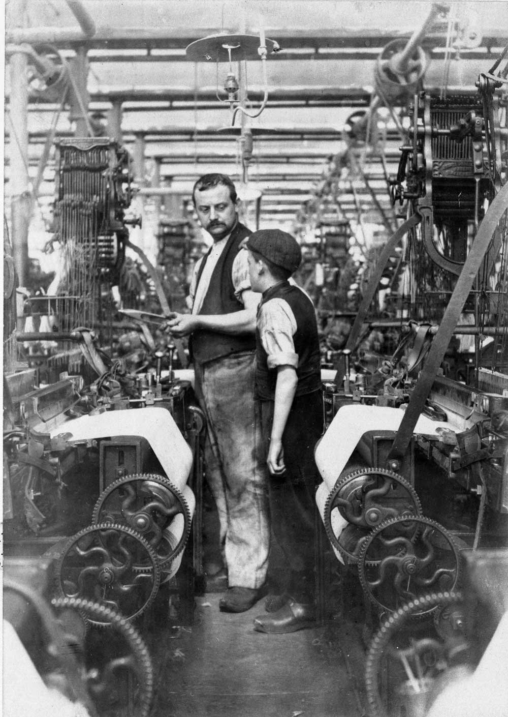 A man in a waistcoat looks down at a boy wearing a flat cap in the aisle between mill equipment.