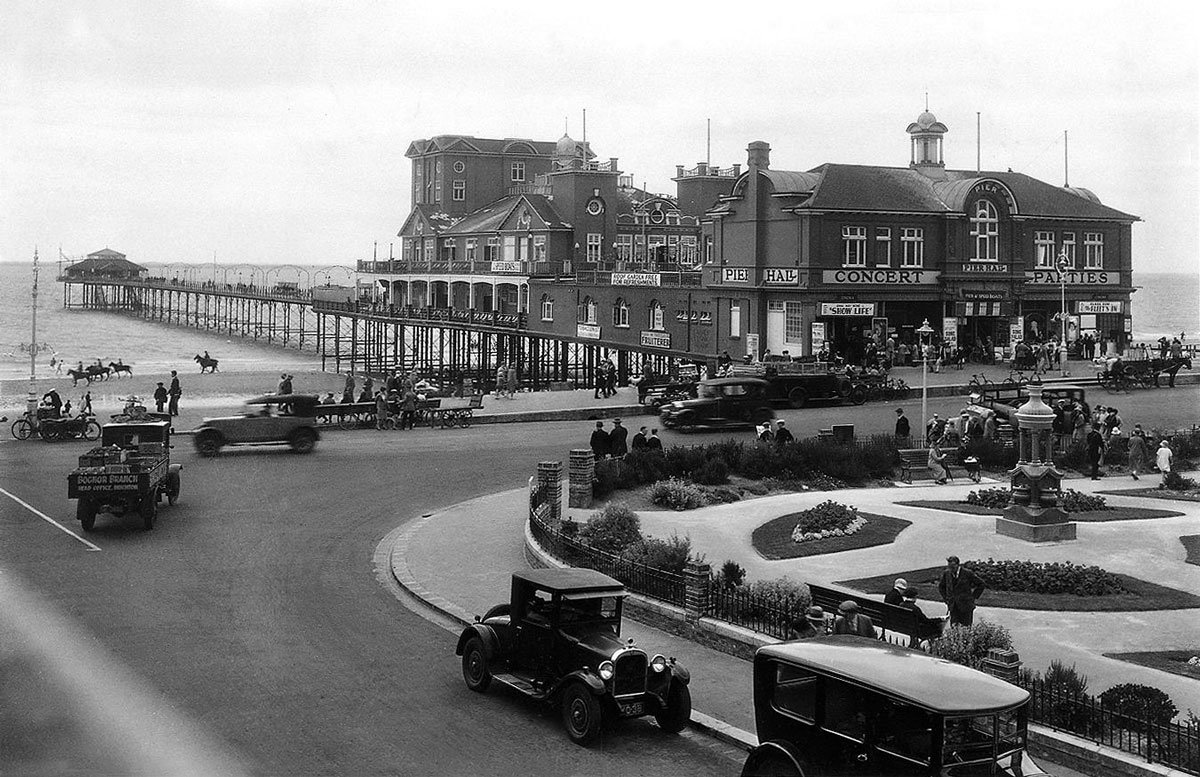 Large buildings sit at the base of a long pier, with cars driving in front.
