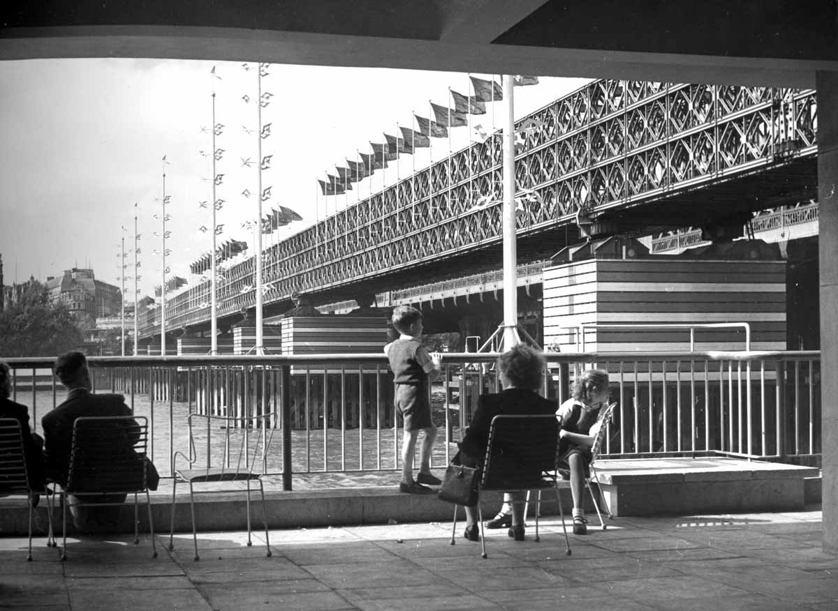A woman sits on a chair with children either side of her, looking across a river by a large bridge..