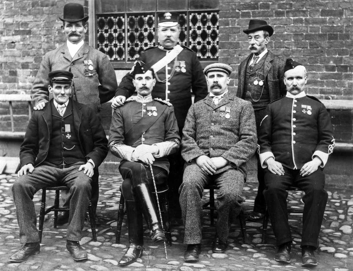 Posed black and white photo of seven men, four on chairs and three standing behind, wearing medals.