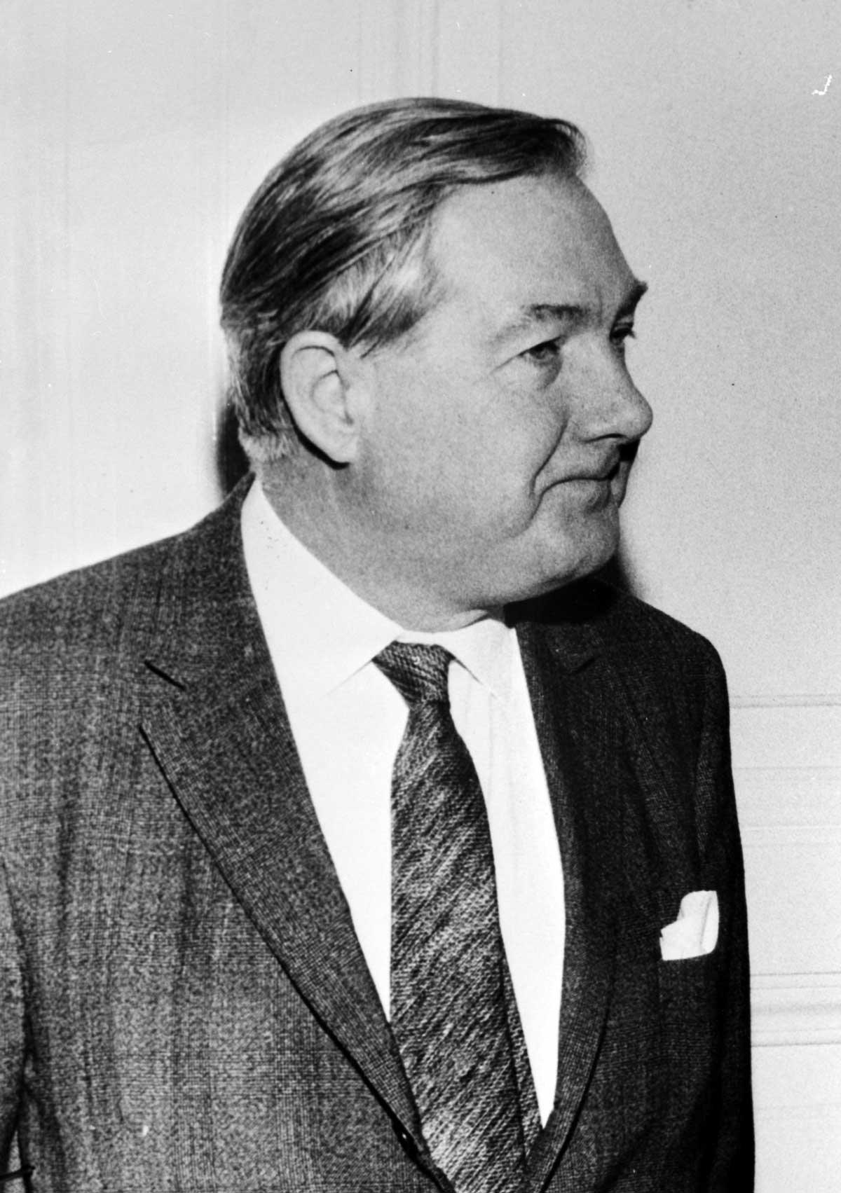 Black and white photo of a man in a dark suit and tie and short hair combed back, pursing his lips.