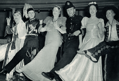 Six partygoers dressed elaborately lock arms and smile as they kick a leg up in unison.