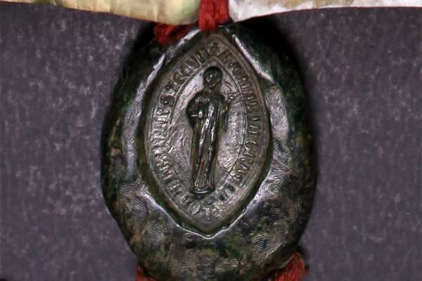 A dark, mottled, oval-shaped seal showing a human figure surrounded by words attached to red string.