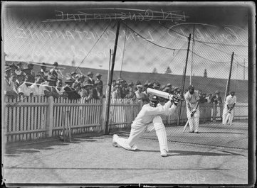 A young black man in cricketing whites photographed in the middle of hitting the ball. A crowd watch