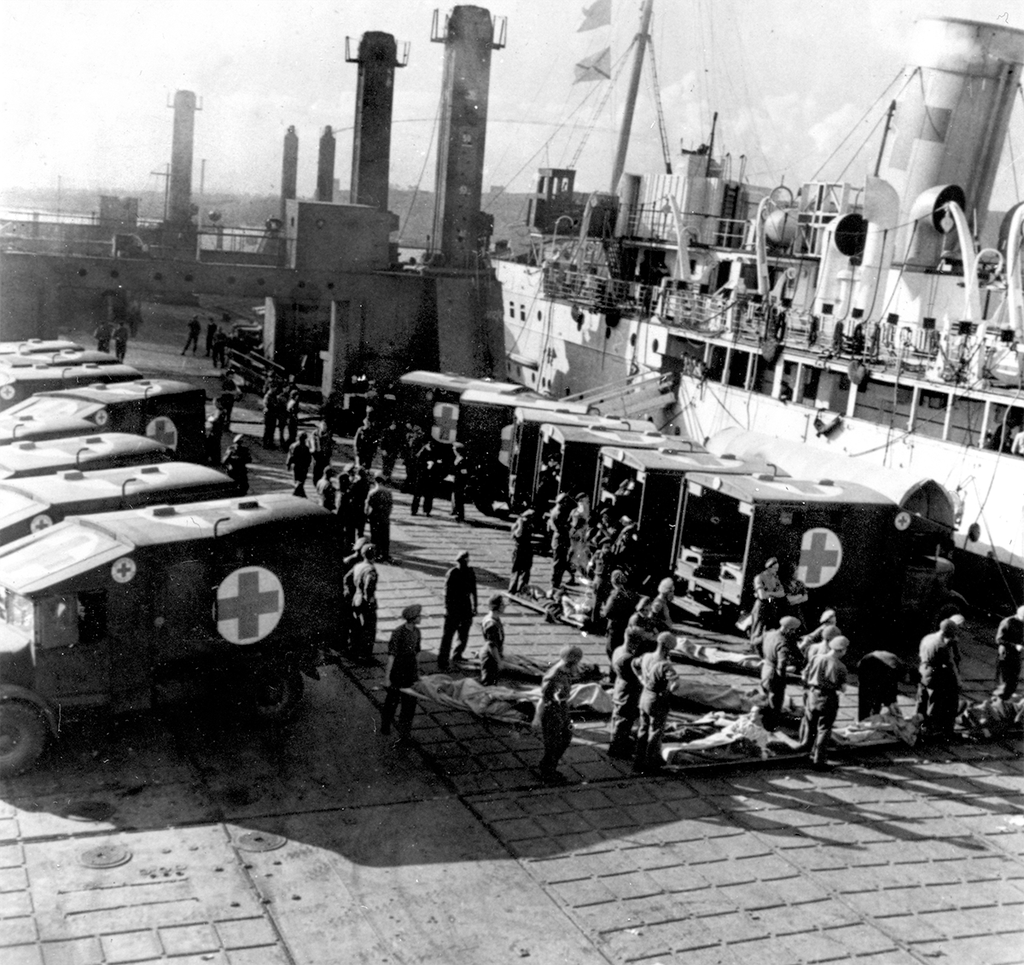 Photograph looking down on around thirty people unloading stretchers from vans marked with crosses.