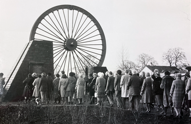 A large wheel from a coal pit is above ground as a memorial. Tens of people are walking towards it.