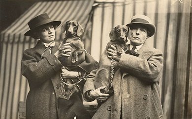 Two women in overcoats and hats hold up dachshunds to the camera
