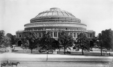Black and white photo of the Royal Albert Hall, a grand domed building, behind some trees.