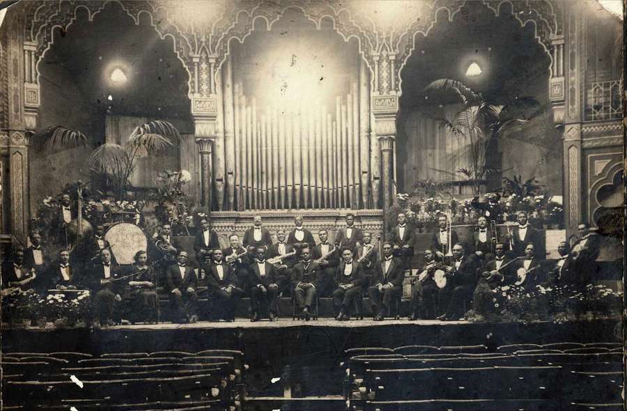 Several dozen, smartly-dressed musicians holding instruments on a highly-decorated stage.