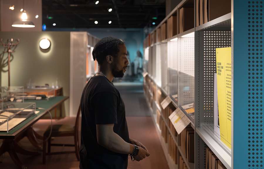 A man gazes at a display case while standing in front of a period office desk.