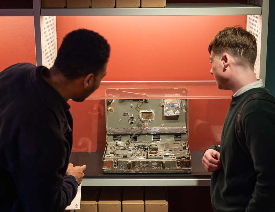 Two people stood looking at a rusted metal case covered with dials, buttons and vents.